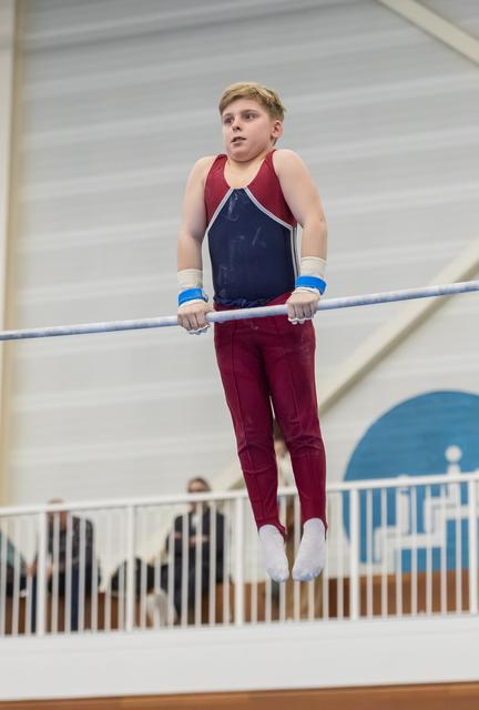 Young gymnast in maroon and navy leotard grips horizontal bar with focused expression during training routine