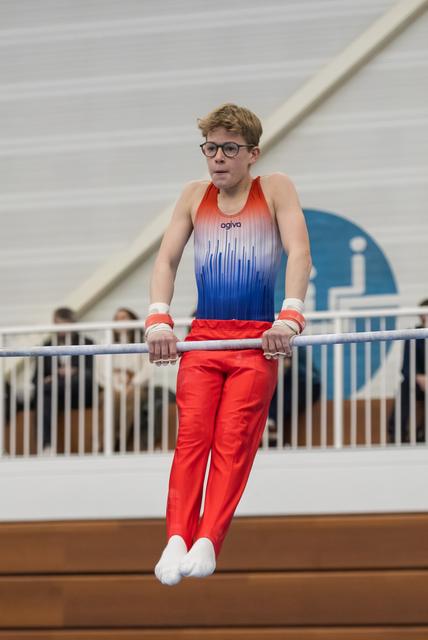 Young gymnast in red and blue gradient uniform concentrates while performing on horizontal bar, wearing glasses