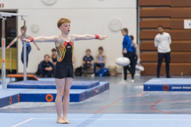 Young male gymnast in black leotard extends arms during floor routine, showing focused concentration in gymnasium