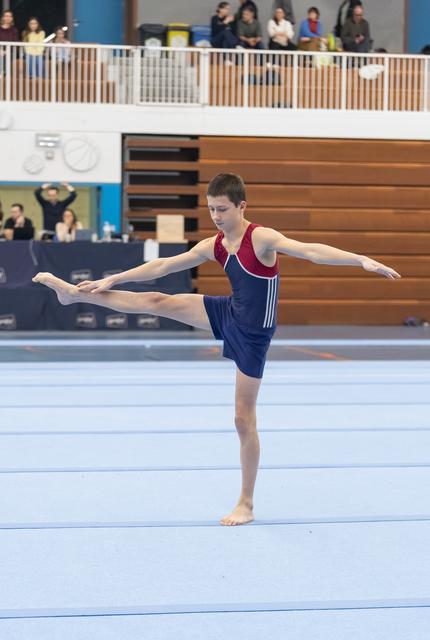 Young gymnast performs floor routine with extended leg pose, showing focus and balance in red and navy leotard