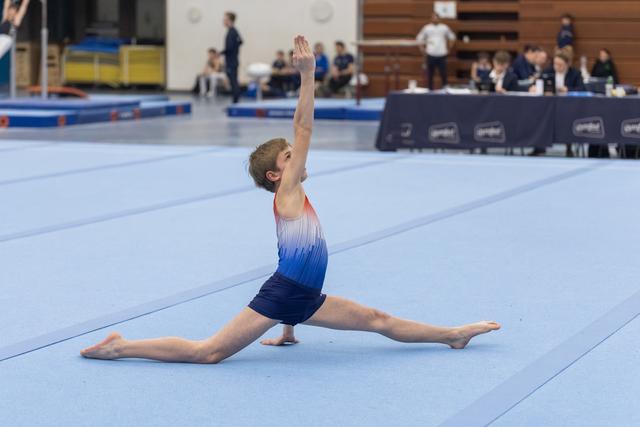 Young gymnast performs a graceful split with raised arm during floor routine on blue mat at indoor gymnastics meet