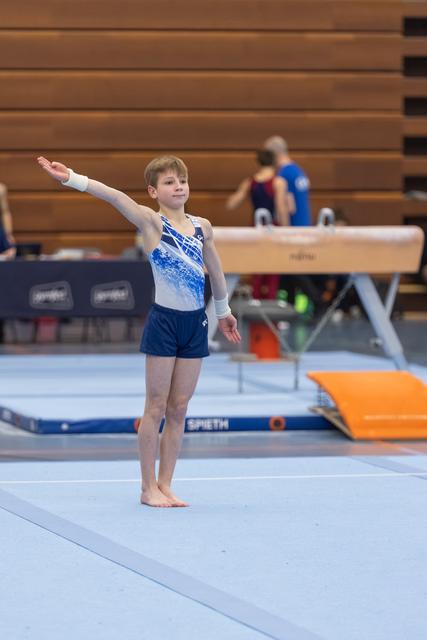 Young gymnast performing floor routine with one arm extended, focused expression, blue leotard, gymnasium setting