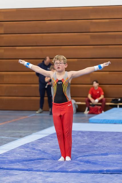 Young gymnast in red pants and black leotard performs floor routine with arms extended, showing focused concentration