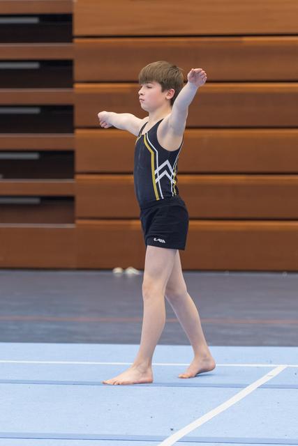 Young gymnast performs floor routine with arms extended, demonstrating focused form on blue mat in gymnasium