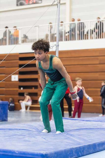 Young gymnast in teal uniform performs floor routine with focused expression in gymnasium with spectators in bleachers