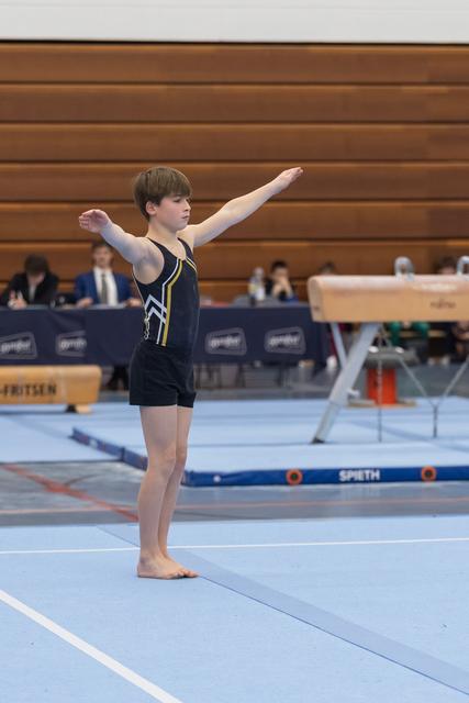 Young gymnast in black leotard stands in starting position on floor mat, arms extended in preparatory pose before routine