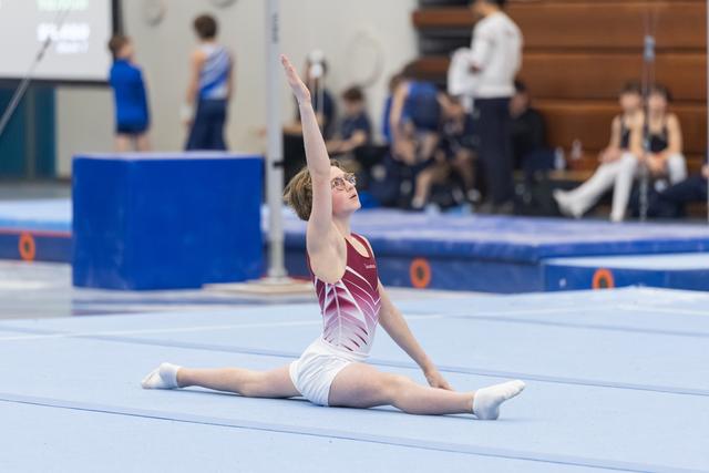 Young gymnast performs a split on floor exercise, arms extended gracefully upward during her routine at indoor meet.