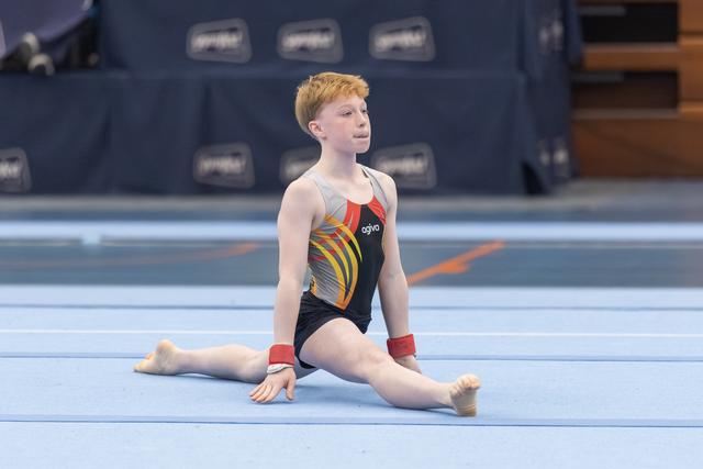 Young gymnast in colorful leotard performs a split position on the floor mat, looking to the side with focused expression
