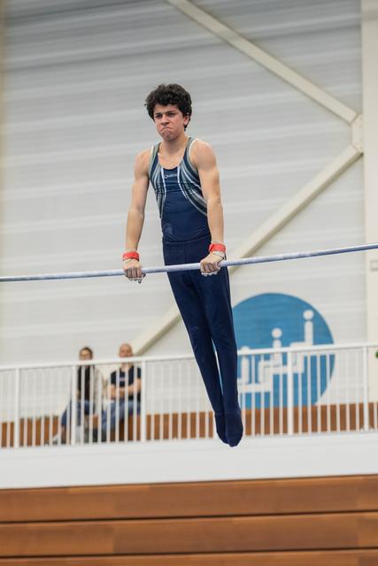 Male gymnast in navy uniform performs horizontal bar routine with intense focus, spectators visible in background