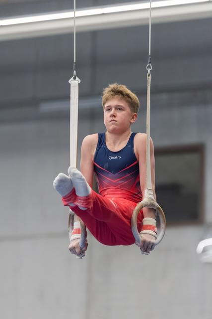Young male gymnast performing on rings with focused upward gaze, wearing red and navy leotard in indoor training facility