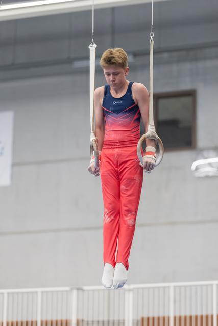 Young male gymnast performs on rings, displaying intense concentration while suspended in the air in gradient outfit