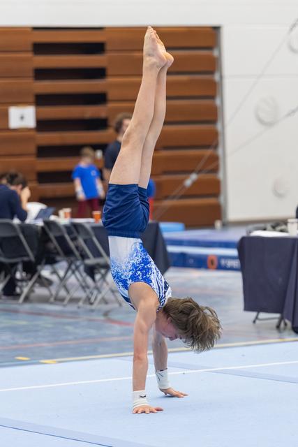 Young gymnast performs a handstand on blue floor exercise mat, legs extended vertically, while spectators watch from bleachers