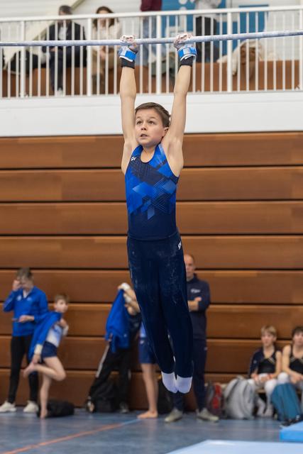 Young gymnast in blue leotard hangs from high bar with focused expression during routine in gymnasium