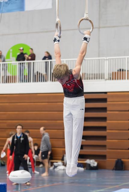 Young gymnast in white pants and burgundy leotard hangs from still rings with extended arms during routine