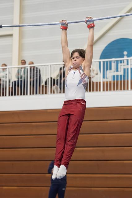 Young gymnast hangs from high bar with focused expression, wearing white leotard and maroon pants in indoor training facility