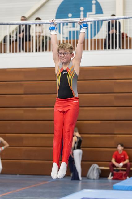 Young gymnast in red pants and colorful leotard hanging from horizontal bar, arms raised, determined expression behind glasses