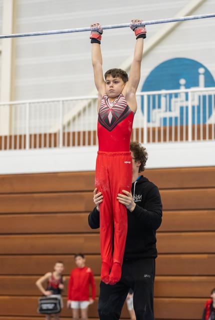 Young gymnast in red outfit hangs from horizontal bar with focused expression while coach spots from below