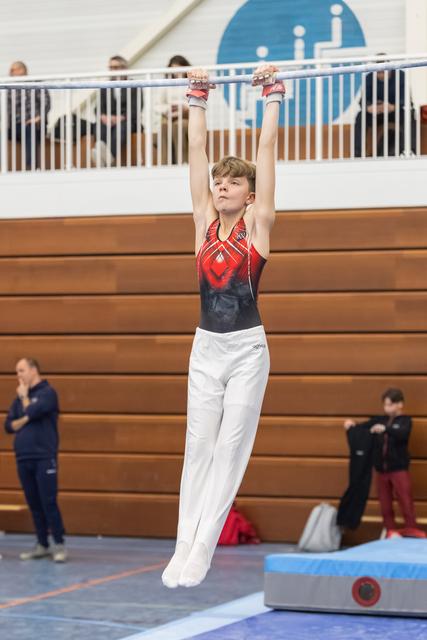 Young gymnast in red and black leotard hangs from horizontal bar with focused expression during routine