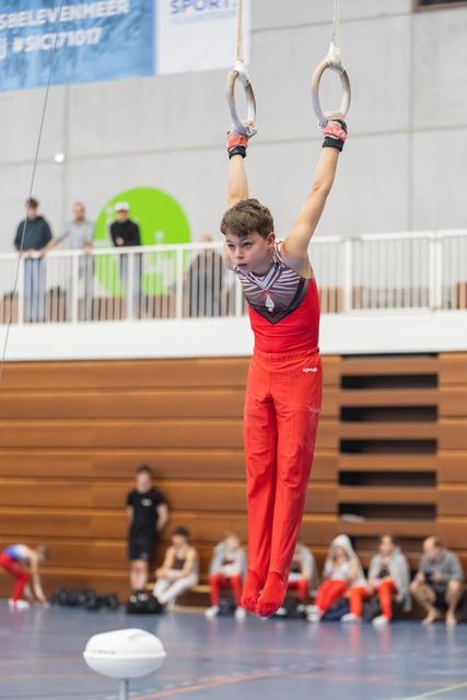 Young gymnast in red outfit hangs from rings with focused expression, arms extended overhead in indoor training facility
