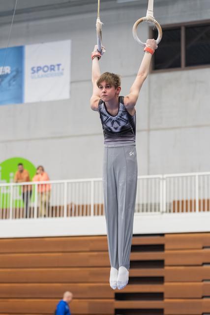 Young male gymnast in gray attire hanging from still rings with focused expression during training session