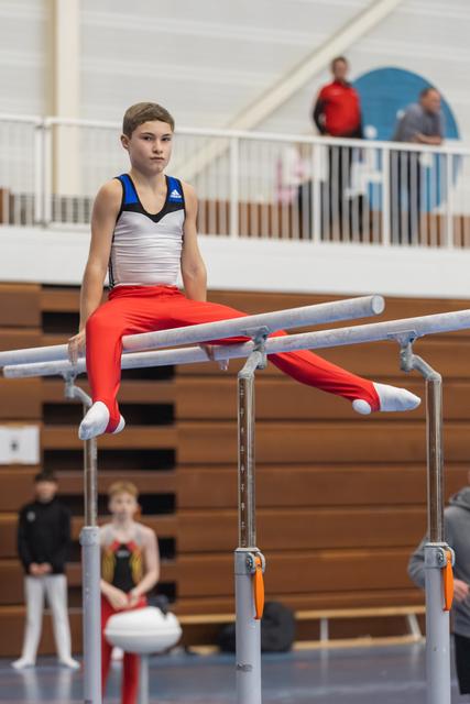 Young gymnast in red pants and white top holds a seated position on parallel bars during training session