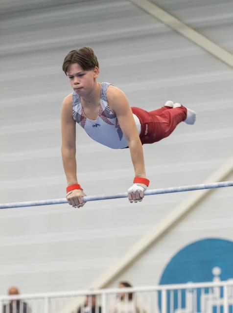 Young gymnast performing horizontal bar routine with focused expression, body parallel to ground in indoor training facility