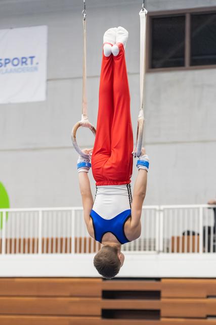 Young gymnast performing an inverted hang on rings, body fully extended upward in red shirt and blue-white shorts