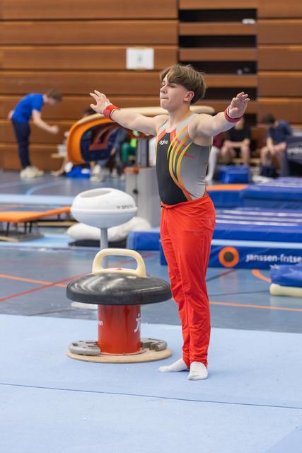 Young gymnast in red pants and gray leotard strikes a pose with arms extended beside a mushroom circle apparatus