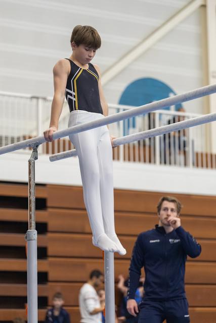 Young gymnast in navy leotard performs on parallel bars with focused concentration while coach observes from bleachers