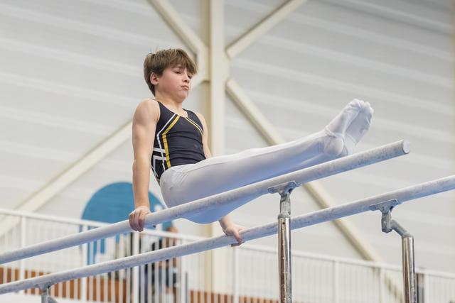 Young gymnast performs L-sit position on parallel bars with focused expression, legs extended horizontally in white pants and navy leotard