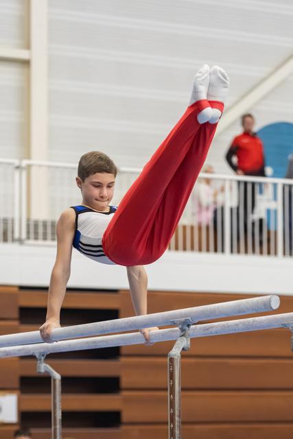 Young gymnast performs L-sit hold on parallel bars wearing red pants and white leotard, focused expression during routine