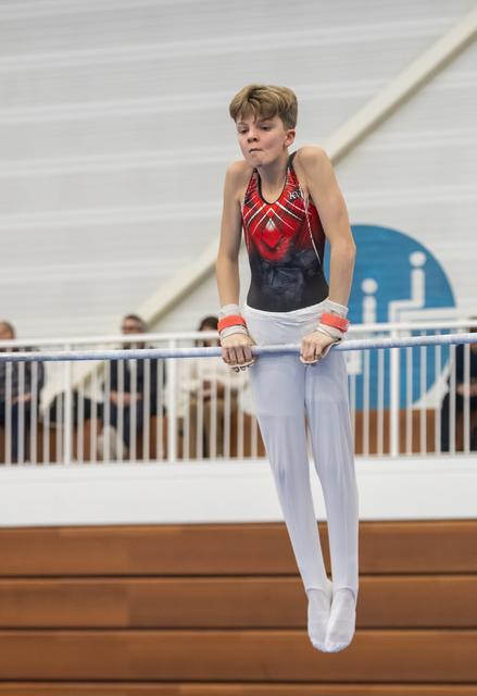 Young gymnast in red and navy leotard performs on horizontal bar, displaying focus and concentration during routine
