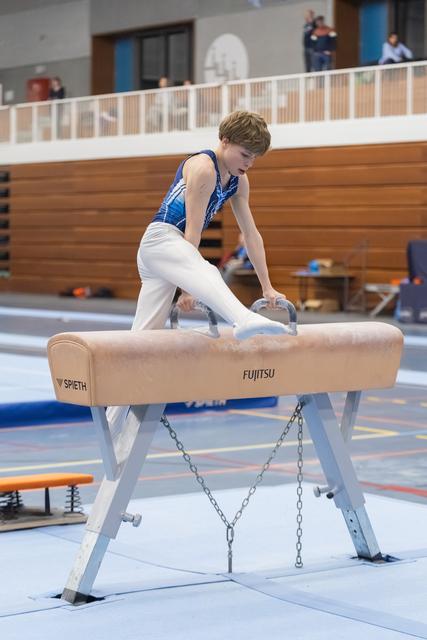 Young male gymnast in blue and white uniform performs skill on pommel horse during indoor gymnastics meet