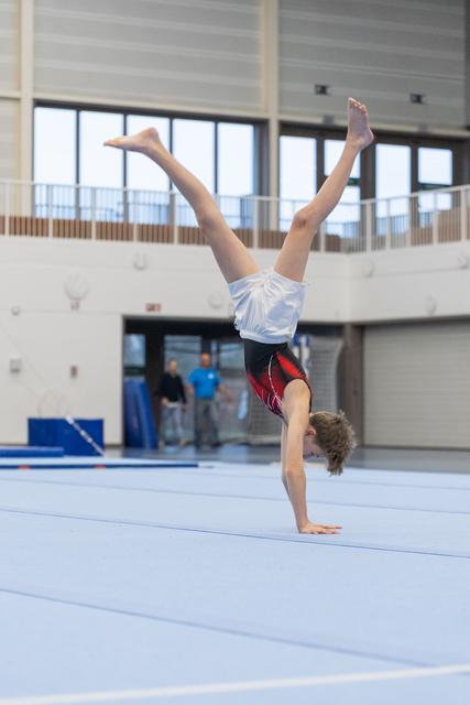 Young gymnast executing a one-armed handstand with legs split during floor exercise at indoor gymnastics facility