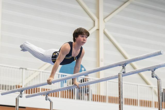 Young gymnast holds himself on parallel bars with extended legs during routine, displaying focused concentration