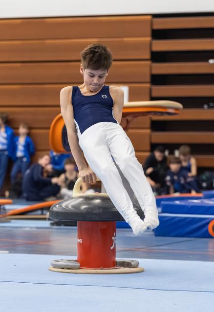 Young gymnast in navy and white uniform performs a scissors move on the pommel horse during indoor meet