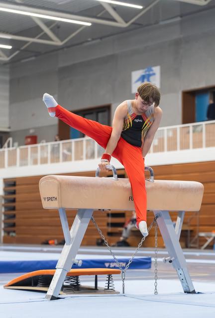 Young gymnast in orange outfit performs a scissor move on pommel horse, displaying strength and concentration in training hall