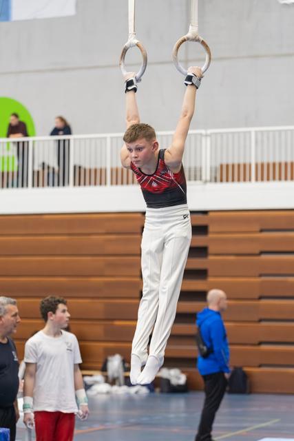 Boy in red plaid leotard hangs from gymnastics rings with focused expression during training at indoor gymnasium