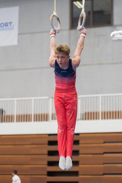 Young male gymnast in red gradient leotard executes a hang position on still rings during training or competition