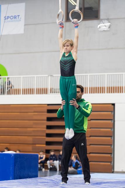 Young gymnast in teal uniform hangs from rings with arms extended while coach spots below in indoor training facility