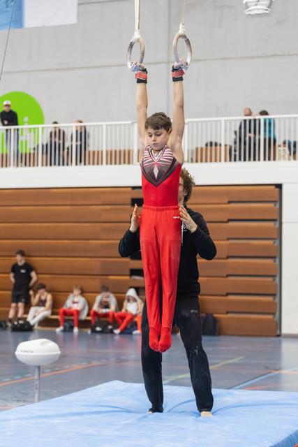 Young gymnast in red uniform performs rings routine with coach supporting from below, arms extended overhead in gymnasium