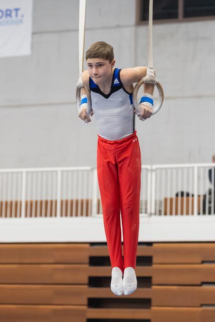 Young male gymnast performing on still rings in white and red uniform, displaying focused concentration during apparatus work
