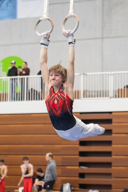 Young gymnast executing an L-sit position on still rings, displaying strength and focus in a gymnasium setting