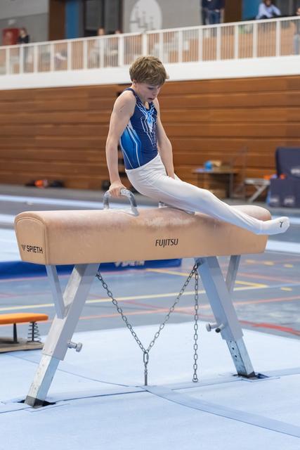 Young gymnast in blue leotard executes scissors skill on pommel horse, demonstrating control and leg extension during training