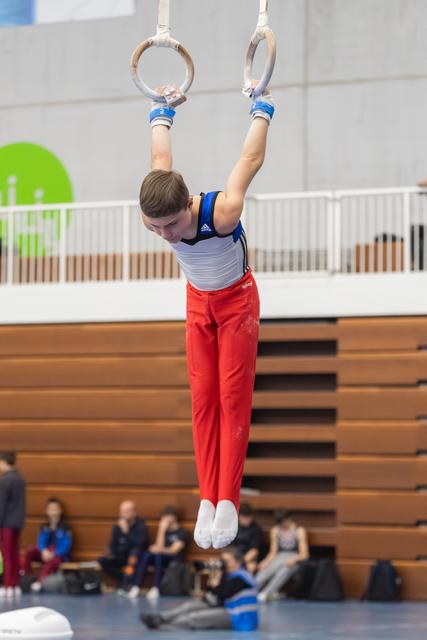 Young gymnast in red pants and white-blue leotard performing a hang position on still rings during indoor meet