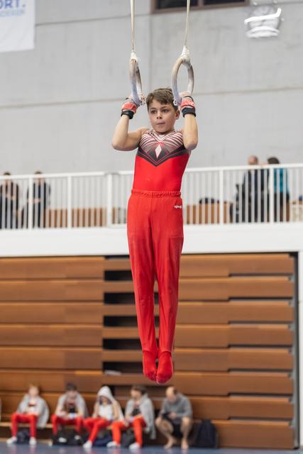 Young gymnast in red uniform holds a steady position on the rings during a routine, displaying focus and strength