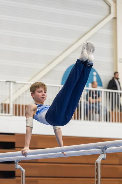 Young gymnast in blue leotard demonstrates an inverted L-position hold on parallel bars, showing concentration and control