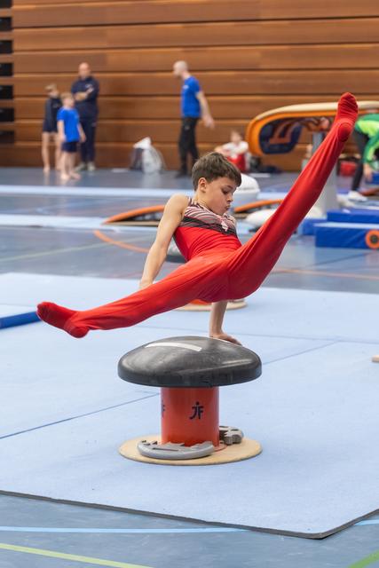 Young gymnast in red performs extended scissor position on pommel horse during training in indoor gymnasium