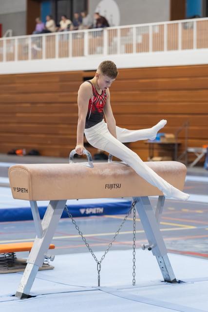Young gymnast performs a scissor movement on pommel horse, legs extended in white pants, focused concentration.