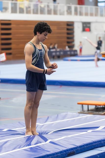 Young male gymnast stands on trampoline, adjusting his hands in preparation, wearing dark singlet in indoor gym facility